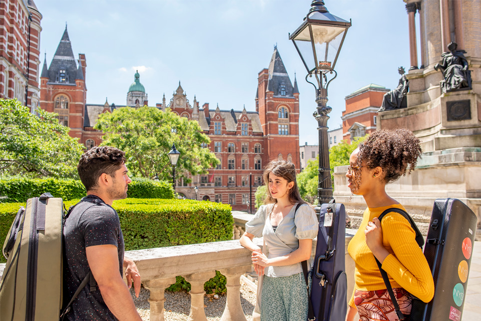 A group of students, carrying instrument cases on their back, chatting to each other, with the Royal College of Music main building in the background.